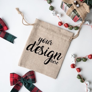 Könnte beinhalten: Ein Geschenksack aus Jute mit Kordelzug. Der Sack ist mit dem Text "your design here" in schwarzer Schrift verziert. Der Sack ist von roten und grünen karierten Schleifen und einer Girlande aus Glöckchen umgeben.
