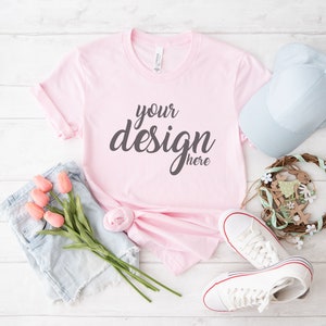 Op de afbeelding: Een roze T-shirt met de tekst "your design here" in grijs handschrift. Het T-shirt ligt op een wit houten oppervlak met een blauwe spijkerbroek, een lichtblauwe baseballpet, een paar witte sneakers en een kleine bloemenkrans.