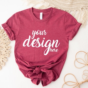 Puede incluir: Una camiseta de color granate con el texto "your design here" en letras blancas. La camiseta está anudada en la parte inferior y colocada sobre una superficie blanca.