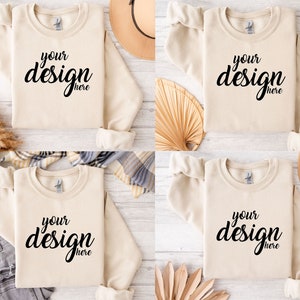 Pu&ograve; includere: Quattro felpe beige con la scritta "your design here" stampata sul davanti. Le felpe sono disposte su una superficie di legno bianco con vari oggetti decorativi intorno.