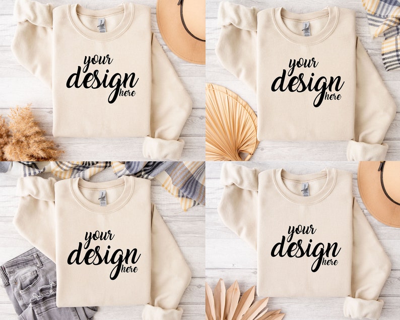 Pu&ograve; includere: Quattro felpe beige con la scritta "your design here" stampata sul davanti. Le felpe sono disposte su una superficie di legno bianco con vari oggetti decorativi intorno.