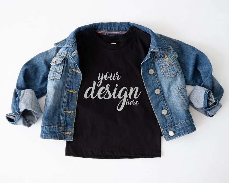 May include: A black t-shirt with the text "your design here" in a white, cursive font. The t-shirt is laying on a white surface with a blue denim jacket on top of it.