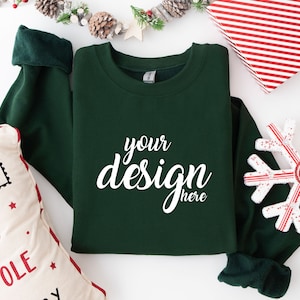 May include: A dark green sweatshirt with the text "your design here" in white on the front. The sweatshirt is laying flat on a white surface with a snowflake ornament and a pillow with a Christmas theme.