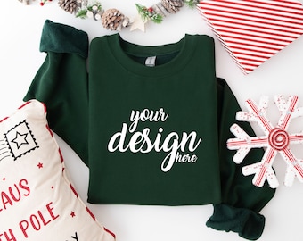 May include: A dark green sweatshirt with the text "your design here" in white on the front. The sweatshirt is laying flat on a white surface with a snowflake ornament and a pillow with a Christmas theme.