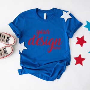 K&ouml;nnte beinhalten: Ein k&ouml;nigsblaues T-Shirt mit dem Text "your design here" in roten Buchstaben. Das T-Shirt liegt flach auf einer wei&szlig;en Oberfl&auml;che mit roten, wei&szlig;en und blauen Sternen, die um es herum verstreut sind.