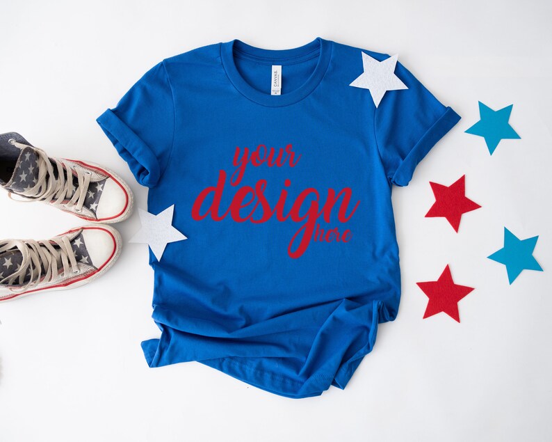 K&ouml;nnte beinhalten: Ein k&ouml;nigsblaues T-Shirt mit dem Text "your design here" in roten Buchstaben. Das T-Shirt liegt flach auf einer wei&szlig;en Oberfl&auml;che mit roten, wei&szlig;en und blauen Sternen, die um es herum verstreut sind.