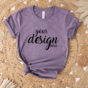 May include: A heathered purple t-shirt with rolled-up sleeves, featuring the text "your design here" in black script. The shirt is knotted at the bottom and rests on a woven jute rug with a natural, neutral color palette. Dried palm leaves add a decorative touch.