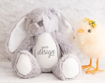 May include: A plush gray bunny with long ears and a white belly sits next to a fluffy yellow chick wearing a floral crown. The bunny has the text "your design here" on its belly. The chick has orange legs.