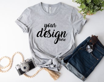 May include: A gray t-shirt with the text "your design here" in black. The shirt is tied in a knot and is laying on a white surface with a pair of denim shorts, sunglasses, and a wooden beaded necklace.