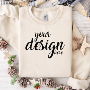 May include: A beige sweatshirt with the text "your design here" on the front. The sweatshirt is laid flat on a white wooden surface with a pine cone garland and a wooden star.