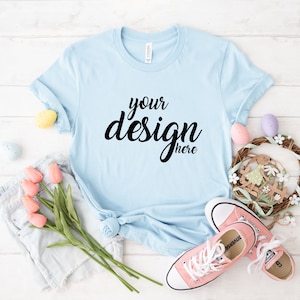 Op de afbeelding: Een lichtblauw t-shirt met de tekst "your design here" in zwart. Het shirt ligt plat op een wit houten oppervlak met een paar roze sneakers, een spijkerbroekje en een bloemenkrans met paaseieren.