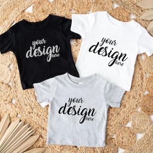 Puede incluir: Tres camisetas en blanco en negro, blanco y gris jaspeado, colocadas sobre una estera de paja tejida. Cada camiseta tiene el texto "your design here" impreso en la parte delantera.