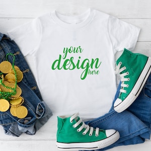 May include: White t-shirt with the text "your design here" in green lettering. The t-shirt is laid out on a white wooden surface with a pair of green high-top sneakers, a denim jacket, and a gold chain with green beads.