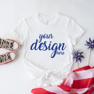 K&ouml;nnte beinhalten: Ein wei&szlig;es T-Shirt mit dem Text "your design here" in blauer Schrift. Das Shirt liegt auf einer wei&szlig;en Oberfl&auml;che mit einer rot-wei&szlig;-blauen amerikanischen Flagge, zwei blauen Windr&auml;dern und einem Paar rot-wei&szlig;-blauer Turnschuhe.