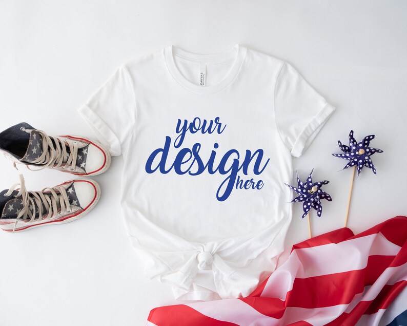 K&ouml;nnte beinhalten: Ein wei&szlig;es T-Shirt mit dem Text "your design here" in blauer Schrift. Das Shirt liegt auf einer wei&szlig;en Oberfl&auml;che mit einer rot-wei&szlig;-blauen amerikanischen Flagge, zwei blauen Windr&auml;dern und einem Paar rot-wei&szlig;-blauer Turnschuhe.