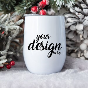 Pode incluir: Um copo térmico de aço inoxidável branco com uma inscrição preta que diz "your design here" sobre um fundo branco nevado e um ramo de abeto congelado com bagas vermelhas.