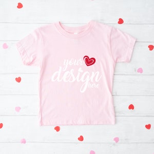 May include: A light pink t-shirt with the words "your design here" in white script, and a red heart graphic. The shirt is surrounded by red and pink heart-shaped confetti on a white wooden surface.