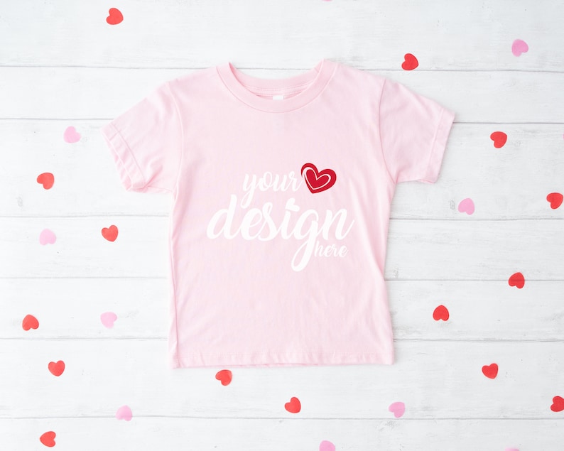 May include: A light pink t-shirt with the words "your design here" in white script, and a red heart graphic. The shirt is surrounded by red and pink heart-shaped confetti on a white wooden surface.