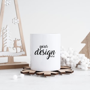 Puede incluir: Taza de cerámica blanca con el texto "your design here" sobre un posavasos de madera con forma de copo de nieve. La taza está rodeada de elementos decorativos blancos y árboles de Navidad de madera.