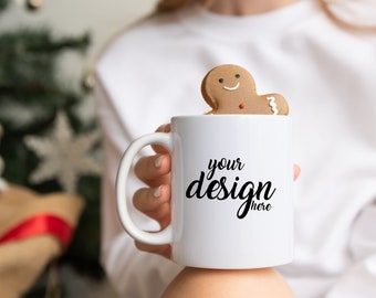 Puede incluir: Una taza de cerámica blanca con el texto "your design here" y una galleta de hombre de jengibre encima. La taza la sostiene una persona. El fondo incluye un árbol de Navidad y decoraciones.