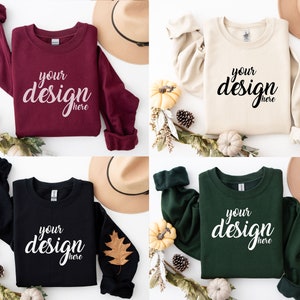 May include: Four different colored sweatshirts with long sleeves, each featuring the text "your design here" in a white, cursive font. The sweatshirts are laid out on a white surface with fall-themed decorations, including pumpkins, pine cones, and leaves.