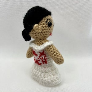 Puede incluir: Muñeca de ganchillo hecha a mano con cabello negro recogido en un moño. La muñeca tiene tez bronceada, ojos negros y labios rojos. Viste un vestido blanco con detalles bordados en rojo.