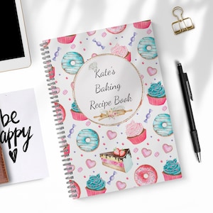 Puede incluir: Un libro de recetas de repostería en espiral blanco con un patrón colorido de donas, cupcakes y caramelos. La portada presenta un círculo dorado con el texto "Kate's Baking Recipe Book".