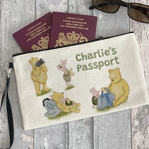 Puede incluir: Una bolsa de lona blanca con cremallera con un tema de Winnie the Pooh. La bolsa tiene una cremallera negra y una correa negra. La bolsa tiene el texto "Charlie's Passport" impreso en ella. La bolsa está decorada con personajes de Winnie the Pooh y Piglet.