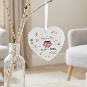 Puede incluir: Un adorno blanco en forma de corazón con el texto "Mrs. Finley" y "Best Tea Cher" en rosa y negro. El adorno está colgado de una cinta blanca y decorado con ilustraciones coloridas. Una taza de té está sobre una mesa de madera.