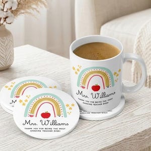 May include: White ceramic mug and coasters with a rainbow and apple design. The mug and coasters have the text "Mrs. Williams, Thank you for being the most amazing teacher ever!" The mug is filled with coffee.