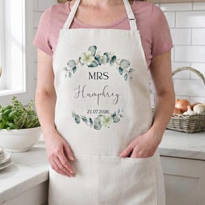 Könnte beinhalten: Eine beigefarbene Küchenschürze mit einem Aquarell-Eukalyptuskranz-Motiv. Der Text lautet MRS Humphrey und das Datum 21.07.2026. Die Schürze hat ein Nackenband, zwei Vordertaschen und wird über einem schlichten malvenfarbenen Kurzarmshirt getragen.