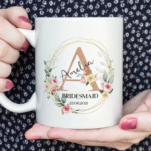 May include: White ceramic mug featuring a floral design with the name "Amelia" and the word "Bridesmaid". The design includes a gold-colored initial "A" and the date "22.06.2021".