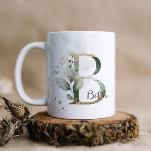 May include: White ceramic mug with a gold and green floral design and the letter "B" in gold. The name "Beth" is written below the letter.