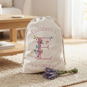 May include: A cream-colored drawstring laundry bag featuring a large pink letter E, the text Elizabeth's LAUNDRY BAG, and several colorful butterfly illustrations. The bag is made of a woven fabric and has thick white rope drawstrings.
