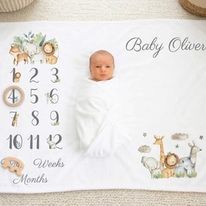 Könnte beinhalten: Weiße Baby-Meilensteindecke mit Illustrationen von Safaritieren, Zahlen und dem Namen "Baby Oliver". Enthält einen Holzring und einen Holzzuschnitt mit dem Text "Tage, Wochen, Monate". Ein Neugeborenes ist in eine weiße Decke gewickelt.