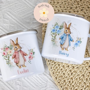Peut inclure: Deux tasses en émail blanc avec des poignées, représentant des illustrations de Lapin de Pâques. Une tasse a un motif floral rose et le nom "Evelyn" imprimé au bas. L'autre tasse a un motif floral bleu et le nom "Henry" imprimé au bas. Les deux tasses ont le texte "Both Side Printed" sur un cercle rose.