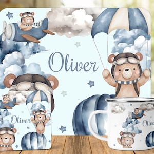 May include: A blue and white personalized baby shower gift set with a bear pilot theme. The set includes a mug, a coaster, and a placemat. The mug and coaster feature a bear pilot flying a plane, while the placemat features a bear pilot parachuting. The set is personalized with the name "Oliver".