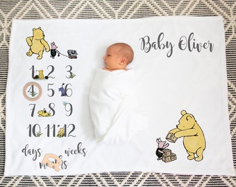 May include: A white milestone blanket featuring Winnie the Pooh and Piglet illustrations, numbers 1 through 12, and the text Baby Oliver. The blanket includes a wooden ring marker for tracking days, weeks, and months, with a swaddled infant lying in the center.