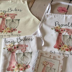 Puede incluir: Dos delantales, una toalla de textura tipo gofre y un cuaderno de espiral con batidoras de pie rosas y motivos florales. Un delantal y la toalla dicen "Tiny Bakes", mientras que el otro delantal y el cuaderno dicen "Pamella". Todos incluyen "Baking is my therapy."