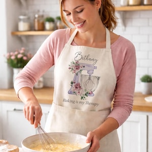May include: A beige apron featuring a printed graphic of a light purple stand mixer adorned with pink and purple floral accents. The text "Bethany" appears above the mixer, and the phrase "Baking is my Therapy" is printed below the illustration.
