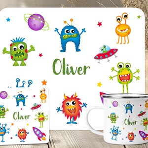 May include: A white enamel mug, coaster, and placemat set featuring colorful cartoon monsters, planets, and rockets. The name "Oliver" is printed on the placemat and coaster in green. The mug has a silver rim and handle. The set is designed for children.