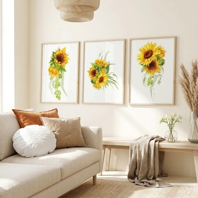 Three framed watercolour art prints hanging side-by-side on a cream wall above a beige sofa. Each print features a vibrant yellow sunflower arrangement with green botanical leaves and trailing vines. The bright, natural light illuminates the modern, cozy living room setting, which includes terracotta and cream cushions, a wooden bench, and a glass vase holding fresh white flowers.