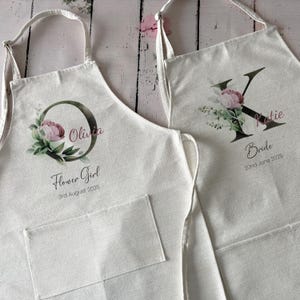 Puede incluir: Dos delantales de color blanco roto con bolsillos delanteros. El delantal izquierdo presenta una letra O grande, el nombre Olivia, el texto Flower Girl y 3rd August 2025. El derecho presenta una letra K grande, el nombre Katie, el texto Bride y 22nd June 2025.