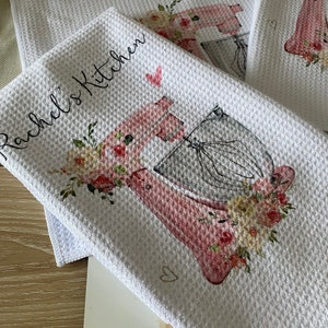 May include: Two white kitchen towels with a pink and grey kitchen mixer design and floral accents. The towels are personalised with the text "Rachel's Kitchen" and "Mami's Kitchen".