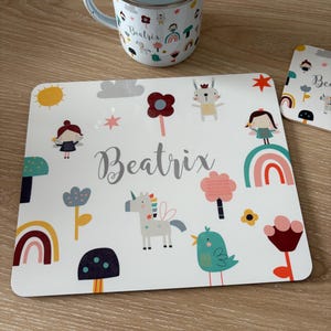 May include: A white placemat and mug set with a whimsical design featuring a unicorn, rainbows, flowers, and the name "Beatrix" in a script font. The mug has a light blue interior and a silver rim.