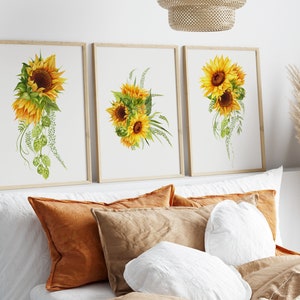 May include: Three framed watercolor paintings of sunflowers with green leaves and stems. The paintings are displayed on a white wall above a bed with pillows.
