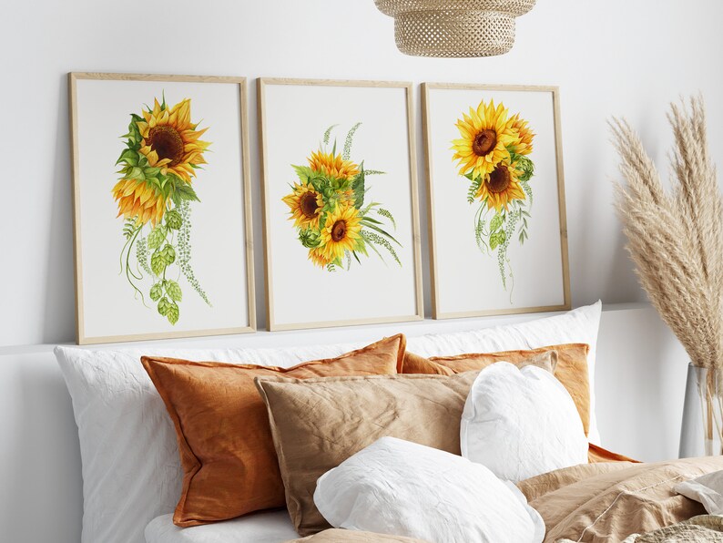 May include: Three framed watercolor paintings of sunflowers with green leaves and stems. The paintings are displayed on a white wall above a bed with pillows.