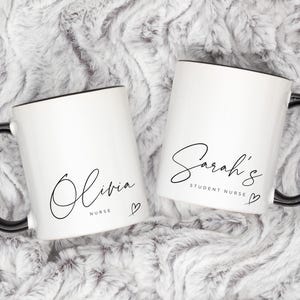May include: Two white ceramic mugs with black handles and black rims. The mugs have black cursive writing on them. One mug says "Olivia Nurse" with a small heart. The other mug says "Sarah's Student Nurse" with a small heart.