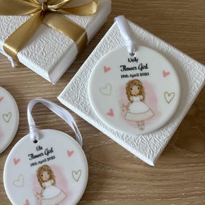 May include: Two white ceramic ornaments with a pink background and a cartoon girl in a white dress holding flowers. The ornaments have gold hearts and the text "Ella Flower Girl 19th April 2025" and "Nelly Flower Girl 19th April 2025".