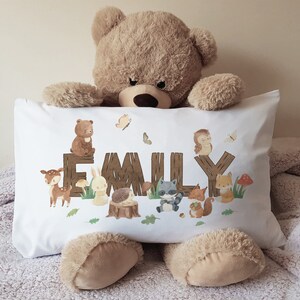 May include: White pillowcase with a woodland animal theme. The pillowcase features a brown bear, a deer, a rabbit, a raccoon, a hedgehog, and a fox. The name "EMILY" is spelled out in brown letters on a wooden fence.